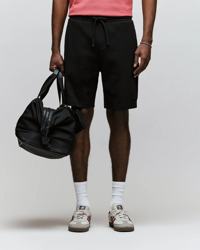 A person stands against a plain light background, wearing Psycho Bunny’s MENS RON WAFFLE CARGO SHORT (MB0900012) in black, paired with a coral waffle knit shirt, white socks, white sneakers with red accents, and holding a black duffel bag.