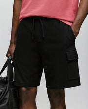 A person is wearing Psycho Bunny’s MENS RON WAFFLE CARGO SHORT - MB0900012 in black, featuring a drawstring, pocket, and subtle embroidered logos, paired with a pink T-shirt and holding a black bag; image is cropped mid-torso to above the knees.