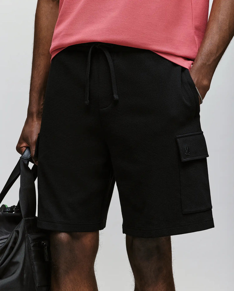 A person is wearing Psycho Bunny’s MENS RON WAFFLE CARGO SHORT - MB0900012 in black, featuring a drawstring, pocket, and subtle embroidered logos, paired with a pink T-shirt and holding a black bag; image is cropped mid-torso to above the knees.