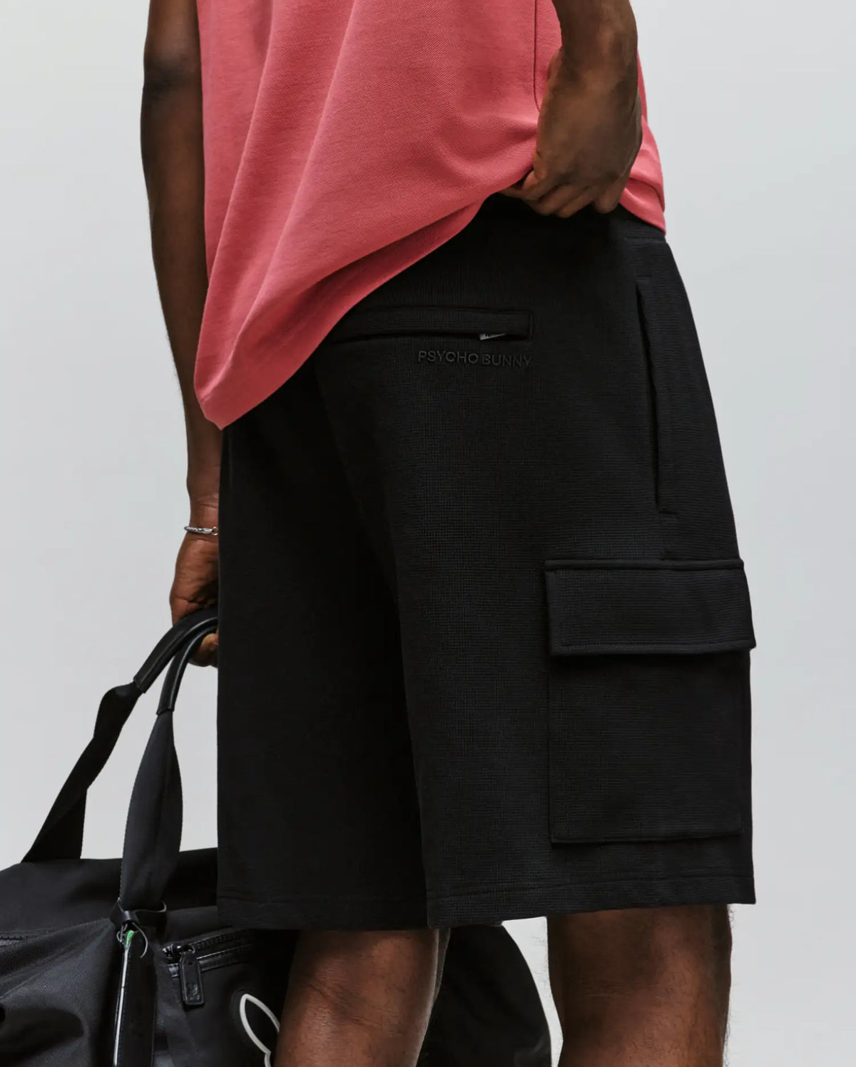 A man in a pink shirt and Psycho Bunny MENS RON WAFFLE CARGO SHORT - MB0900012 with subtle logos and side pockets holds a black duffel bag against a plain, light background.