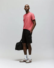 A man confidently poses against a plain background wearing Psycho Bunny’s MENS RON WAFFLE CARGO SHORT - MB0900012, a pink embroidered-logo t-shirt, white socks, sneakers, and carries a black duffel bag with one hand in his pocket.