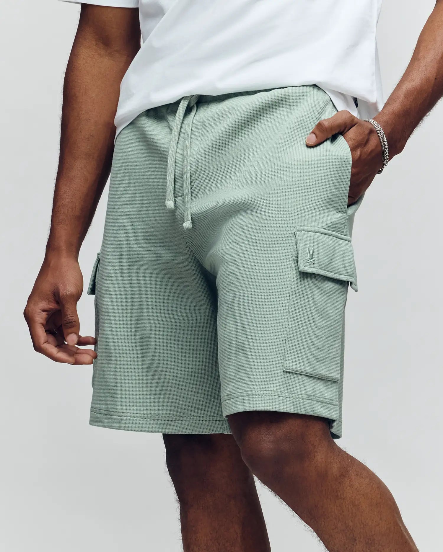 A person wears a white T-shirt and Psycho Bunny MENS RON WAFFLE CARGO SHORT - MB0900012, light green with drawstrings, standing with one hand in a pocket against a plain light background.