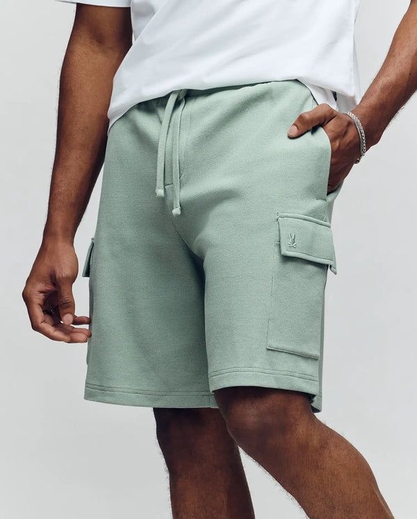 A person wears a white T-shirt and Psycho Bunny MENS RON WAFFLE CARGO SHORT - MB0900012, light green with drawstrings, standing with one hand in a pocket against a plain light background.