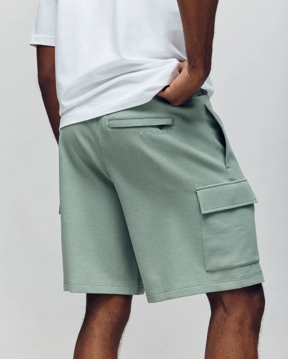 A person is seen from the back wearing Psycho Bunny MENS RON WAFFLE CARGO SHORT - MB0900012 in sage green with flap pockets and visible stitching, paired with a white t-shirt and one hand in their pocket.