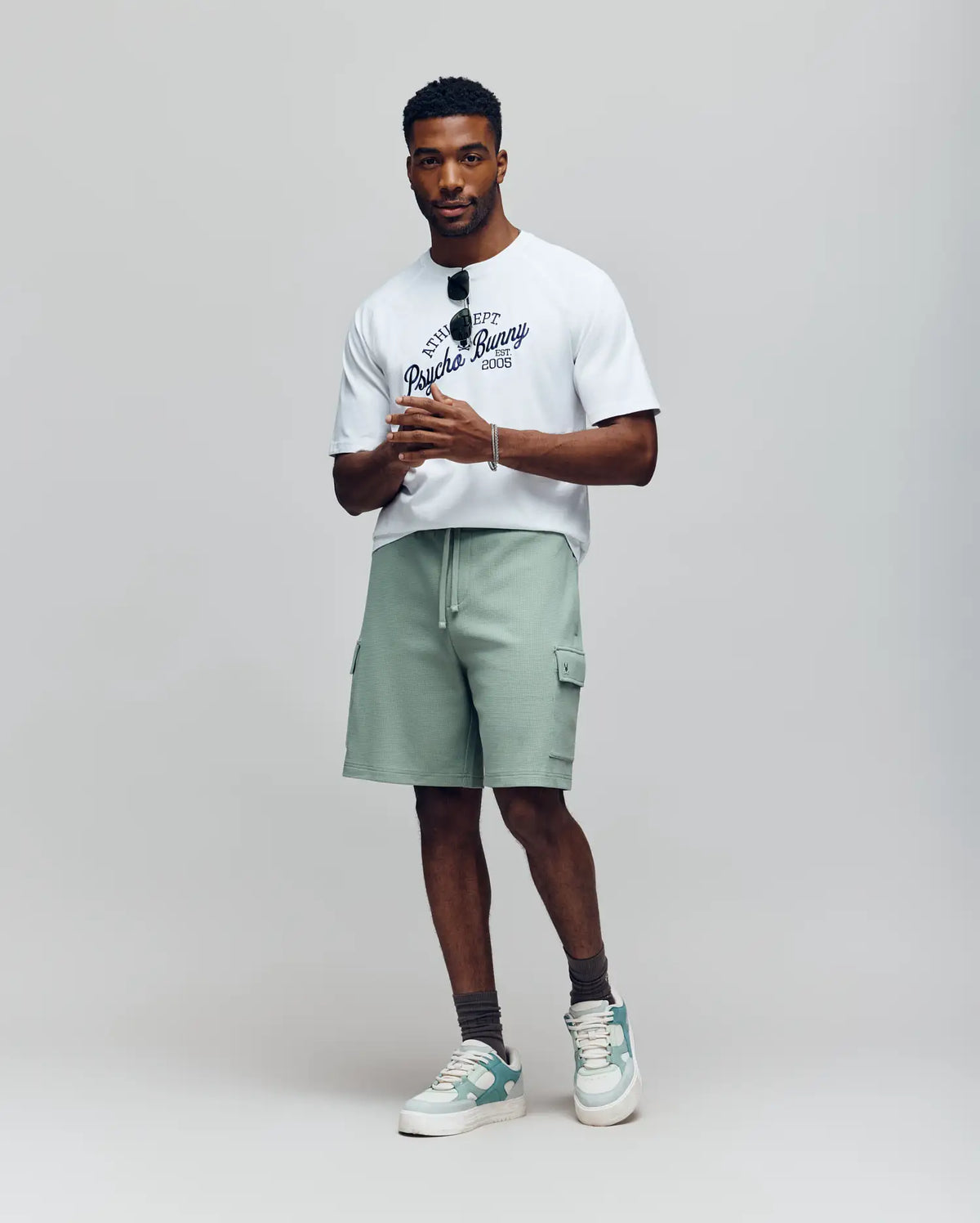 A man stands against a neutral background wearing a white graphic t-shirt, grey socks, pale green and white sneakers, holding sunglasses, and Psycho Bunny MENS RON WAFFLE CARGO SHORT - MB0900012 in light green with drawstring and pockets.