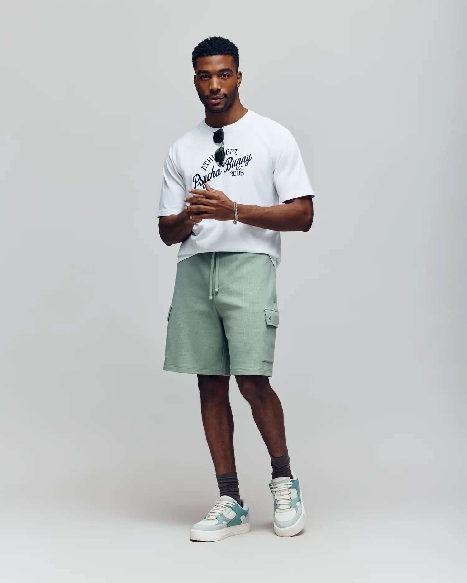 A man stands against a neutral background wearing a white graphic t-shirt, grey socks, pale green and white sneakers, holding sunglasses, and Psycho Bunny MENS RON WAFFLE CARGO SHORT - MB0900012 in light green with drawstring and pockets.