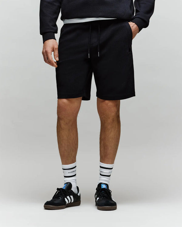 A model showcases the Psycho Bunny MENS KAI SWEATSHORT (MB0900015) with a twill texture, paired with a black sweatshirt, striped white socks, and black Adidas sneakers. The lower half is visible, featuring an embroidered Bunny logo.