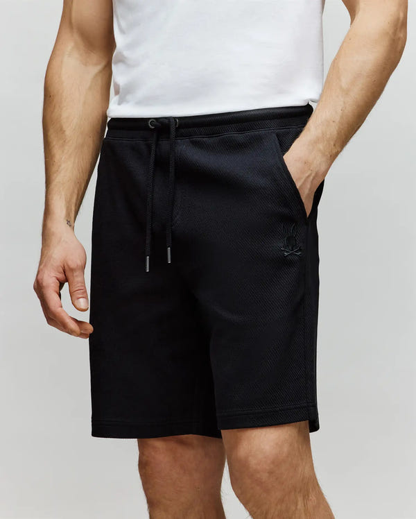 A person wears a white shirt and Psycho Bunny MENS KAI SWEATSHORT - MB0900015, featuring an embroidered Bunny logo and twill-textured fabric, with hands in pockets, shown from shoulders to knees against a plain background.