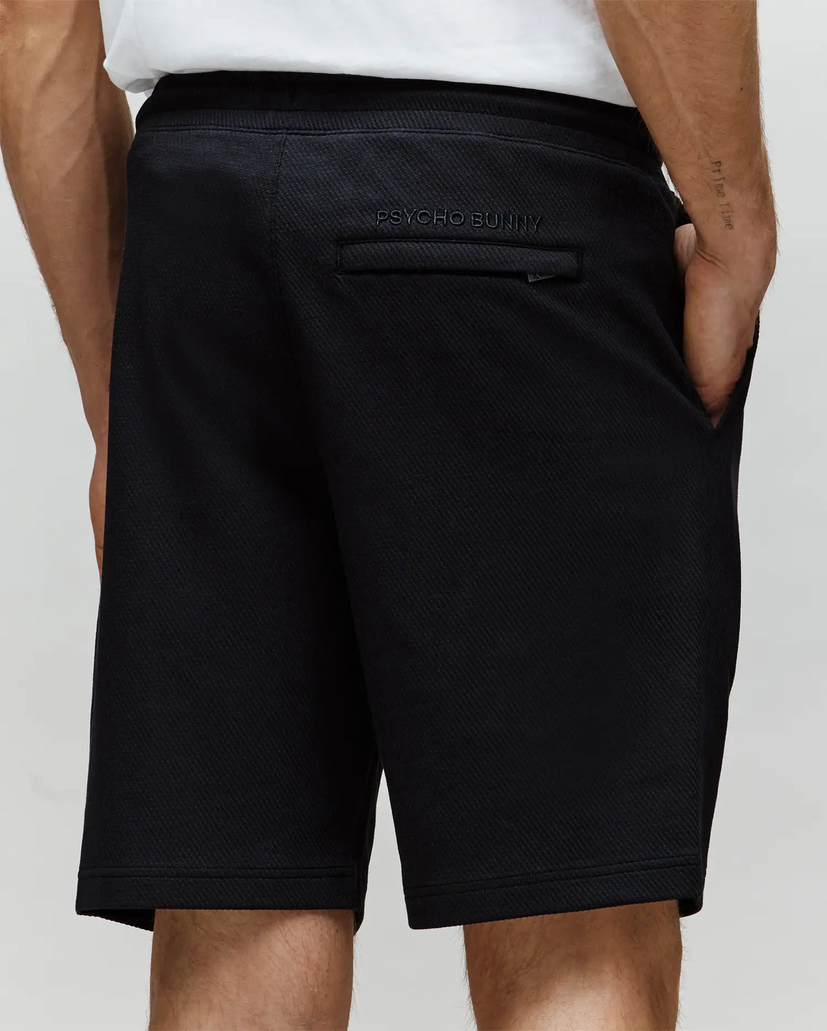 A person wears Psycho Bunny MENS KAI SWEATSHORT - MB0900015 in black with a twill-textured finish, side and single back pocket featuring the embroidered Bunny logo, paired with a white shirt.