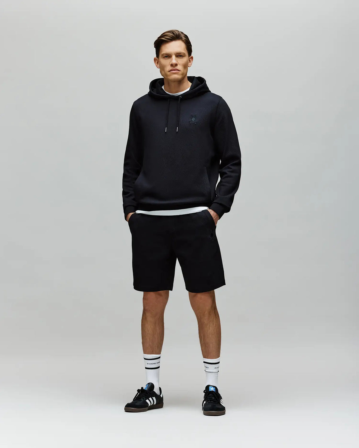 A man stands facing forward against a plain light gray background, wearing a black hoodie, Psycho Bunny MENS KAI SWEATSHORT - MB0900015 with a twill texture, black Adidas sneakers, and white crew socks with black stripes. His hands are in his pockets.