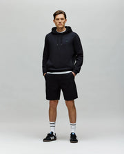 A man stands facing forward against a plain light gray background, wearing a black hoodie, Psycho Bunny MENS KAI SWEATSHORT - MB0900015 with a twill texture, black Adidas sneakers, and white crew socks with black stripes. His hands are in his pockets.