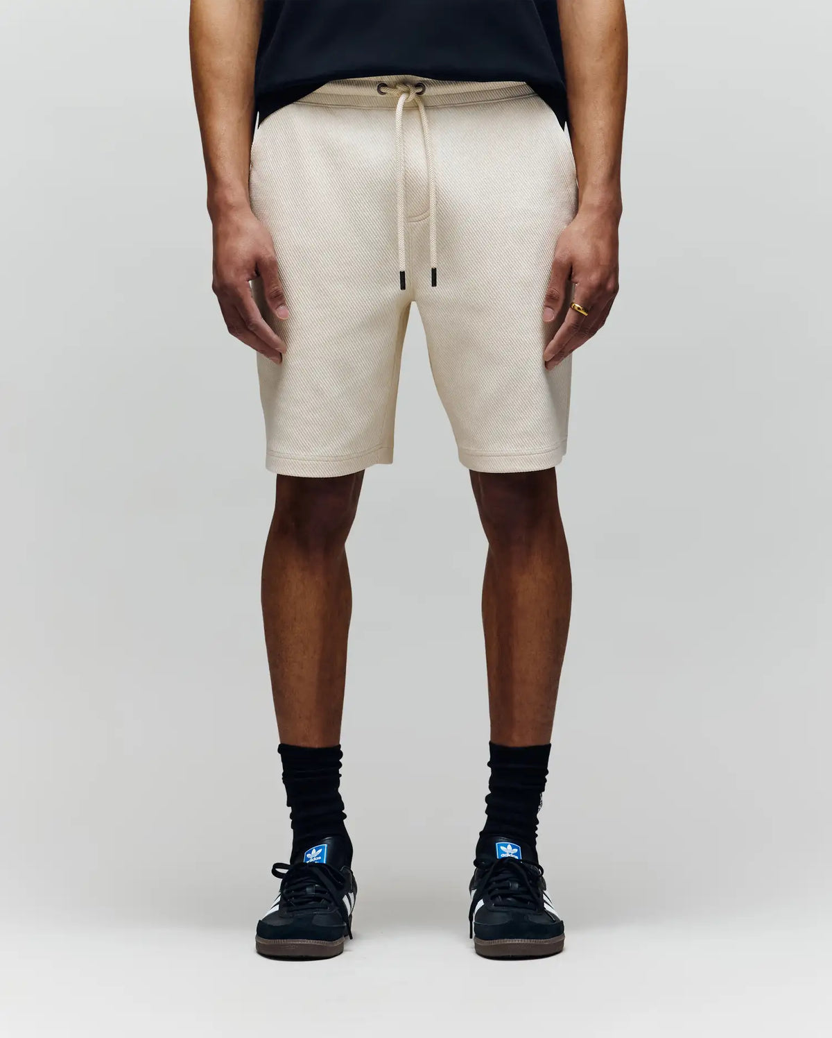 A person wears Psycho Bunny MENS KAI TWILL TEXTURE SWEATSHORT - MB0900015 in beige, featuring a twill texture and embroidered Bunny logo, with black socks, black sneakers, and a black shirt. Only the lower half is visible against a light gray background.
