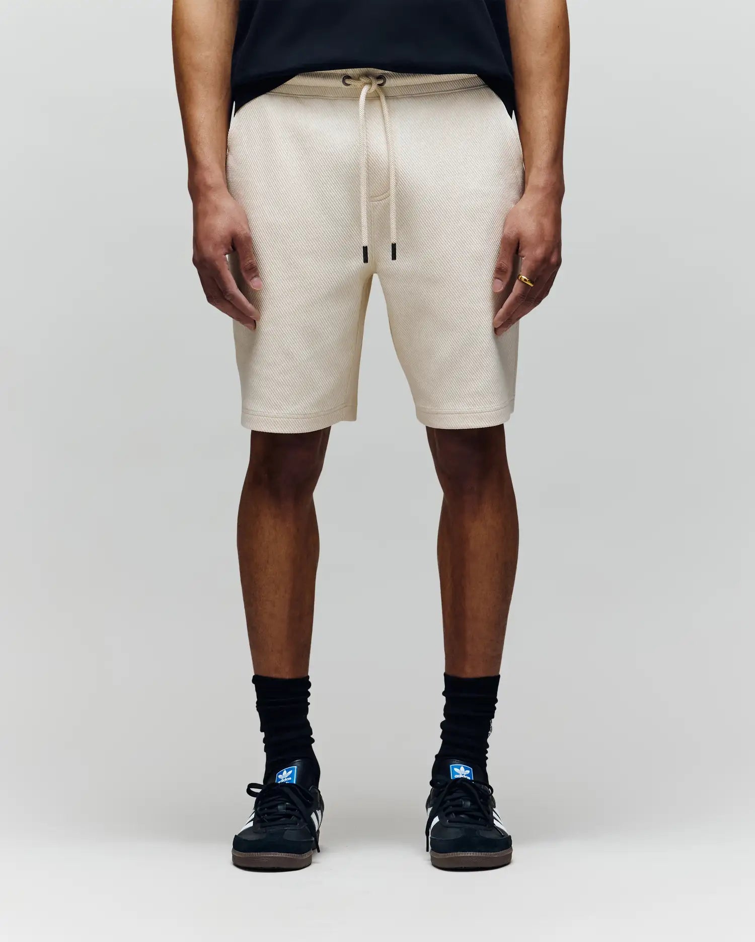 A person wears Psycho Bunny MENS KAI TWILL TEXTURE SWEATSHORT - MB0900015 in beige, featuring a twill texture and embroidered Bunny logo, with black socks, black sneakers, and a black shirt. Only the lower half is visible against a light gray background.