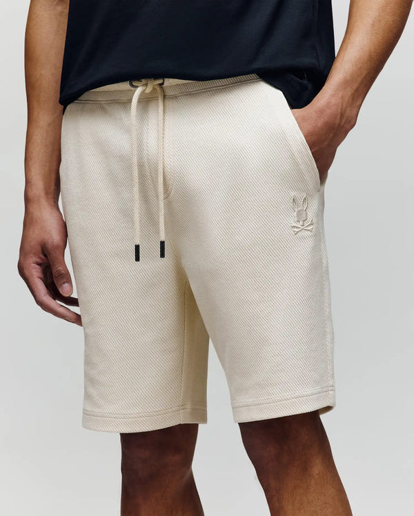 A person wears Psycho Bunny's MENS KAI TWILL TEXTURE SWEATSHORT (MB0900015) in cream, featuring a drawstring waist and embroidered Bunny logo on the left leg, paired with a black t-shirt and their left hand in their pocket.