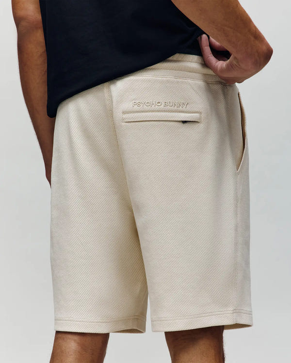 A person, facing away from the camera, wears a black shirt and Psycho Bunny MENS KAI TWILL TEXTURE SWEATSHORT (MB0900015) with a back pocket embroidered with the Bunny logo and "PSYCHO BUNNY." Their right hand is on their hip.