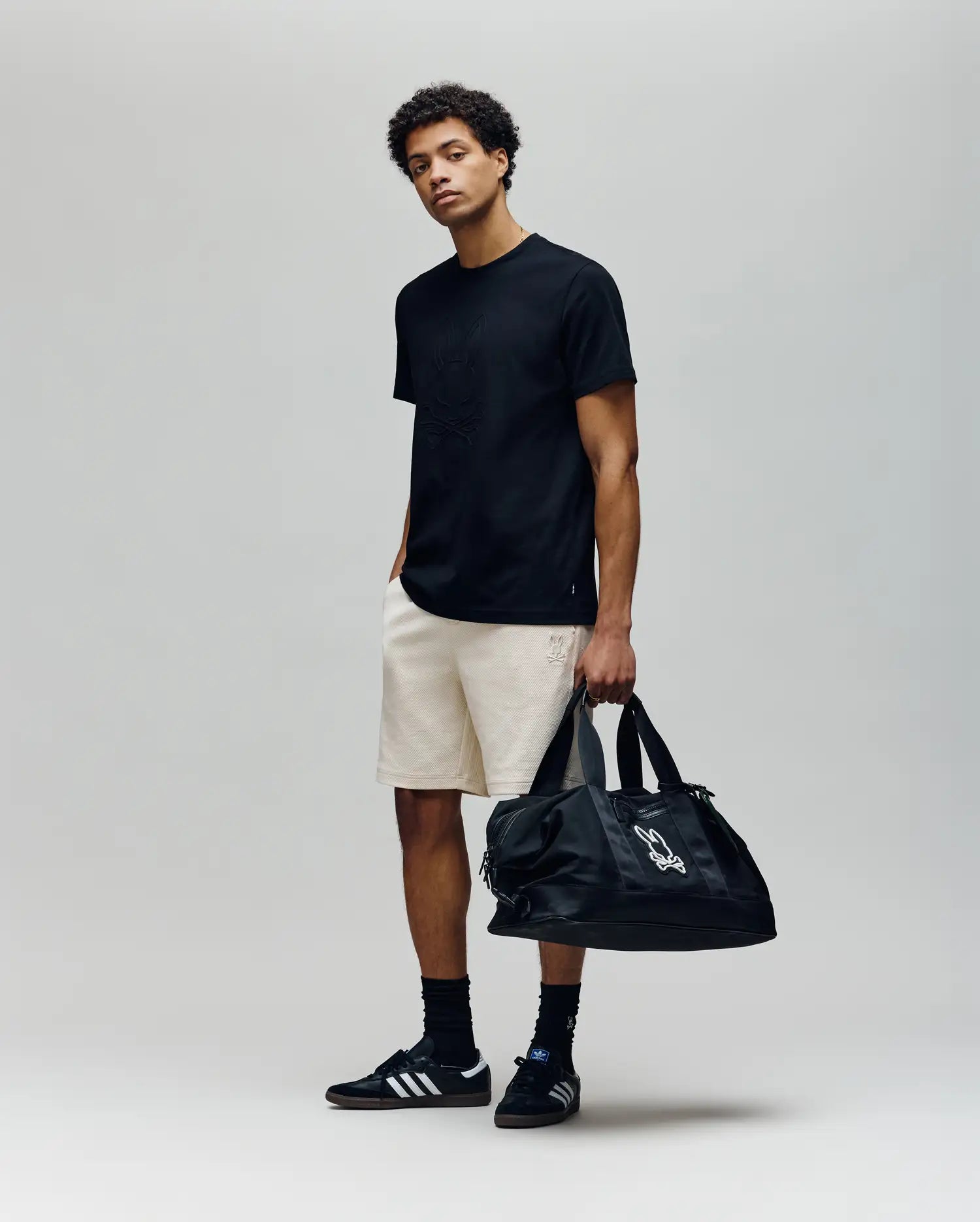 A young man stands against a plain background wearing a black Psycho Bunny t-shirt, MENS KAI TWILL TEXTURE SWEATSHORT - MB0900015, black sneakers and socks. He holds a large black duffel bag in his left hand, right hand in his pocket, looking relaxed.