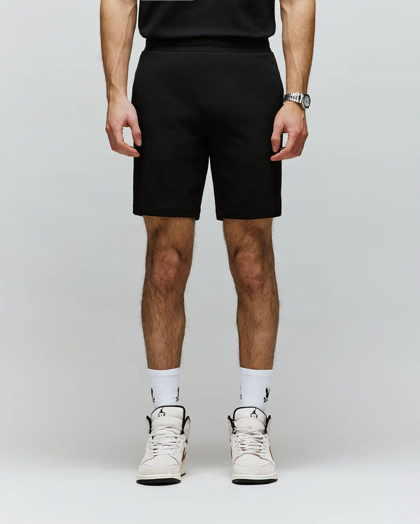 A person, cropped from the shoulders down, wears Psycho Bunny’s MENS LAZSLO SWEATSHORT - MB0900016 in stretch modal-cotton-blend jersey with a black shirt, logo white socks, and white sneakers against a plain light background.