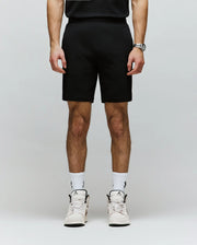 A person, cropped from the shoulders down, wears Psycho Bunny’s MENS LAZSLO SWEATSHORT - MB0900016 in stretch modal-cotton-blend jersey with a black shirt, logo white socks, and white sneakers against a plain light background.