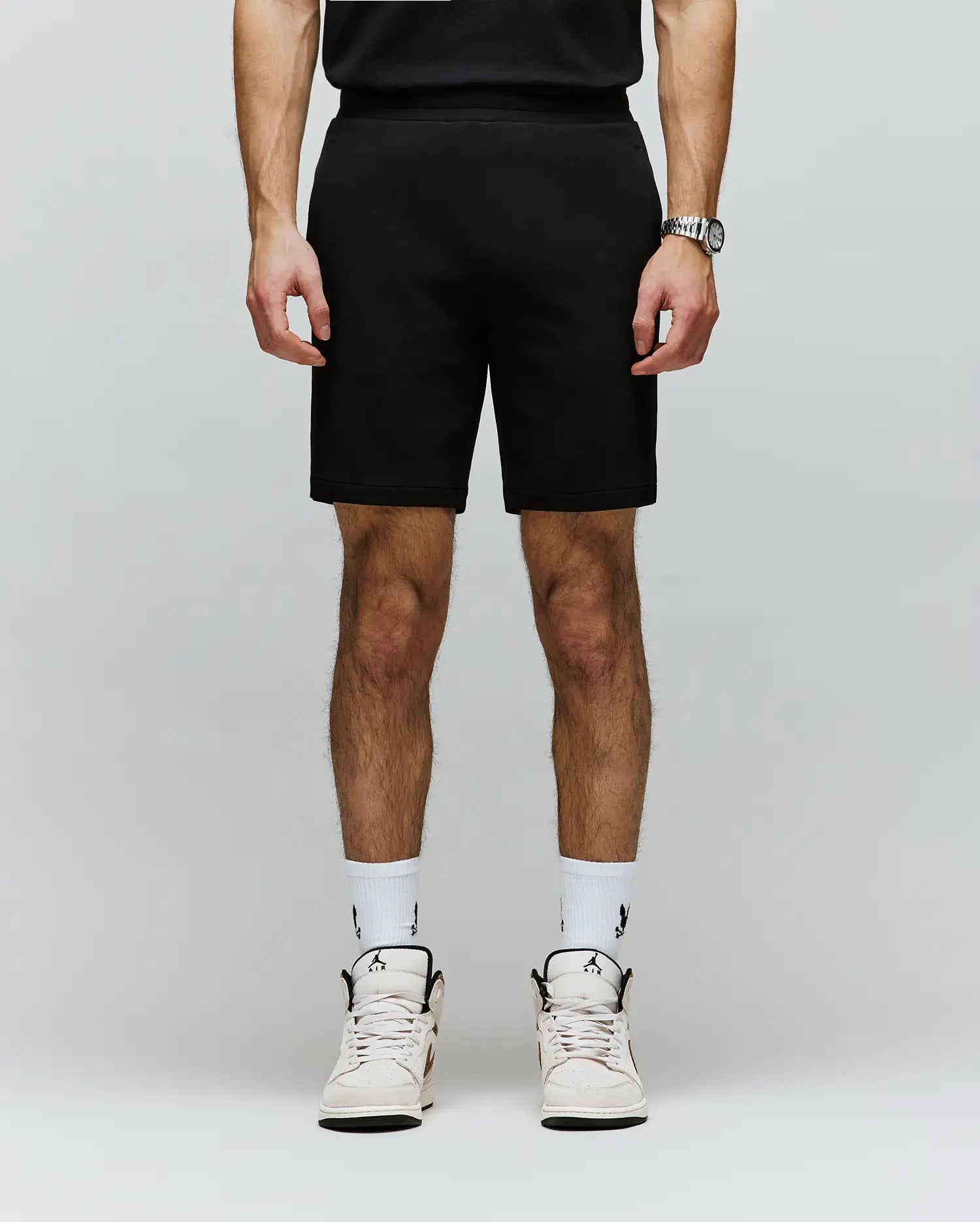 A person, cropped from the shoulders down, wears Psycho Bunny’s MENS LAZSLO SWEATSHORT - MB0900016 in stretch modal-cotton-blend jersey with a black shirt, logo white socks, and white sneakers against a plain light background.