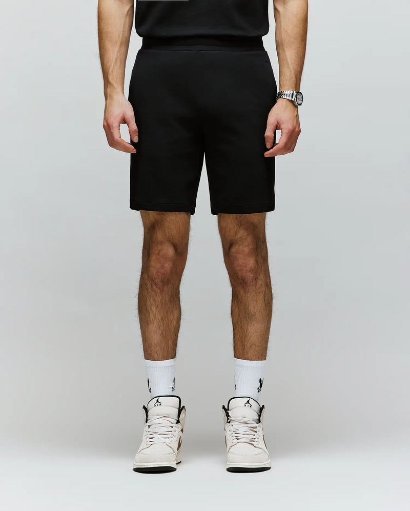 A person, cropped from the shoulders down, wears Psycho Bunny’s MENS LAZSLO SWEATSHORT - MB0900016 in stretch modal-cotton-blend jersey with a black shirt, logo white socks, and white sneakers against a plain light background.