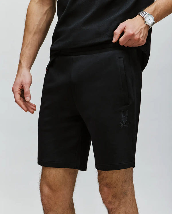 A man wears a black T-shirt with Psycho Bunny’s MENS LAZSLO SWEATSHORT - MB0900016, made from stretch modal-cotton-blend jersey and featuring a small skull and bunny logo on the left thigh. He finishes the look with a silver wristwatch.