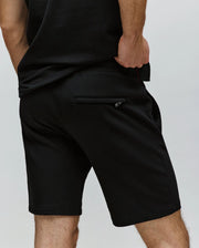 A person stands with one hand in their pocket, wearing Psycho Bunny MENS LAZSLO SWEATSHORT - MB0900016 and a black shirt. The back view showcases the right zippered pocket in comfortable stretch modal-cotton-blend jersey.