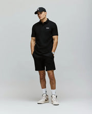 A man stands against a plain background in a black cap, black stretch modal-cotton-blend jersey polo, Psycho Bunny MENS LAZSLO SWEATSHORT - MB0900016, a wristwatch, white socks, and white sneakers with black accents—effortlessly stylish.