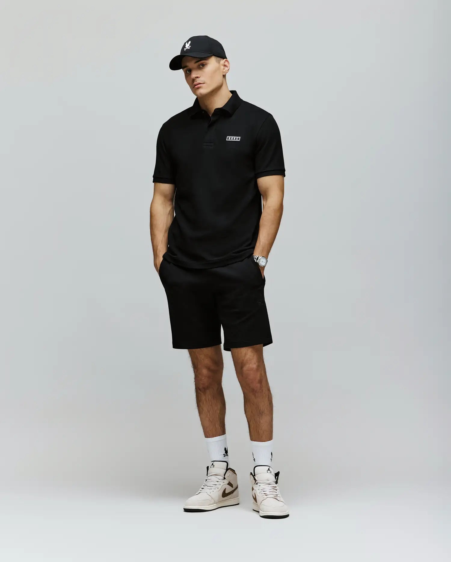 A man stands against a plain background in a black cap, black stretch modal-cotton-blend jersey polo, Psycho Bunny MENS LAZSLO SWEATSHORT - MB0900016, a wristwatch, white socks, and white sneakers with black accents—effortlessly stylish.