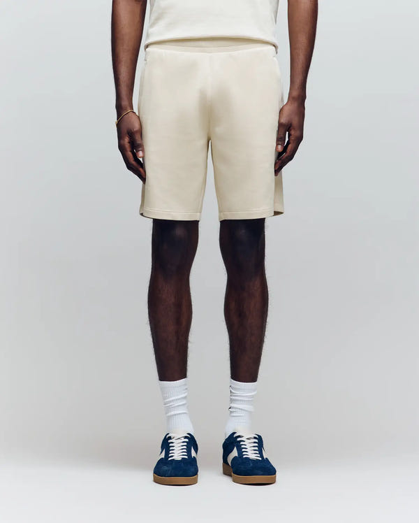 A person wears Psycho Bunny’s MENS LAZSLO SWEATSHORT - MB0900016 in cream, a matching top, white crew socks, and navy sneakers against a light background—an ideal look for effortless casual style.