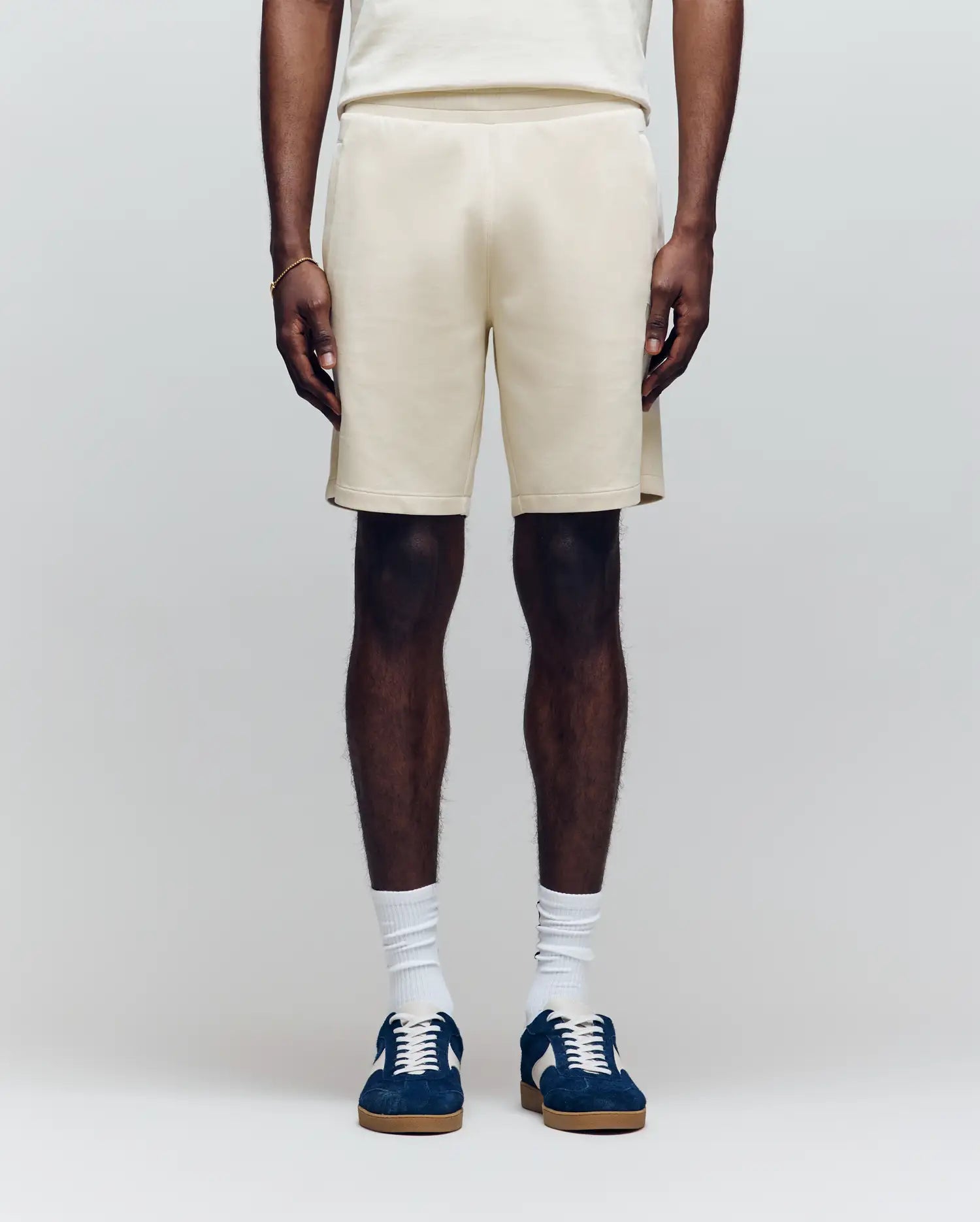 A person wears Psycho Bunny’s MENS LAZSLO SWEATSHORT - MB0900016 in cream, a matching top, white crew socks, and navy sneakers against a light background—an ideal look for effortless casual style.
