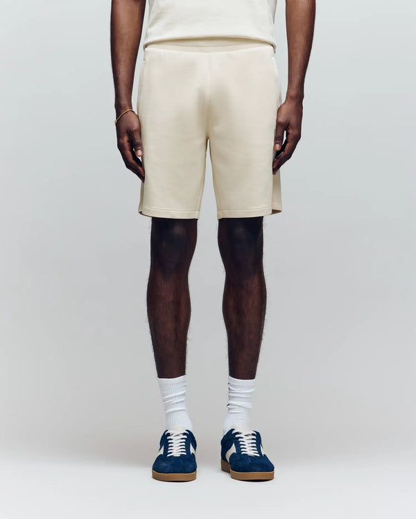 A person wears Psycho Bunny’s MENS LAZSLO SWEATSHORT - MB0900016 in cream, a matching top, white crew socks, and navy sneakers against a light background—an ideal look for effortless casual style.