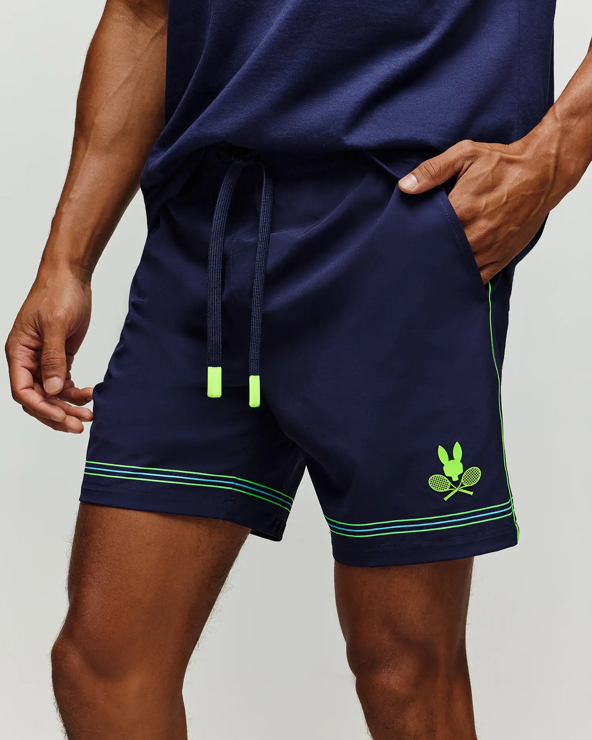 A person wears Psycho Bunny’s TENNIS BALL CREW MENS SHORT (MB0900017) in navy, featuring neon green accents, a drawstring, and a crossed rackets bunny logo graphic on the left leg. Their hand is in the right pocket.