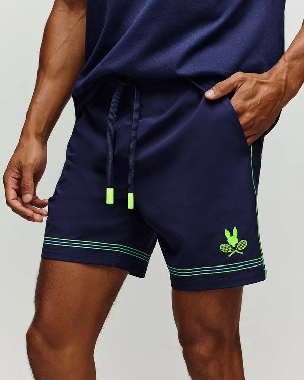 A person wears Psycho Bunny’s TENNIS BALL CREW MENS SHORT (MB0900017) in navy, featuring neon green accents, a drawstring, and a crossed rackets bunny logo graphic on the left leg. Their hand is in the right pocket.