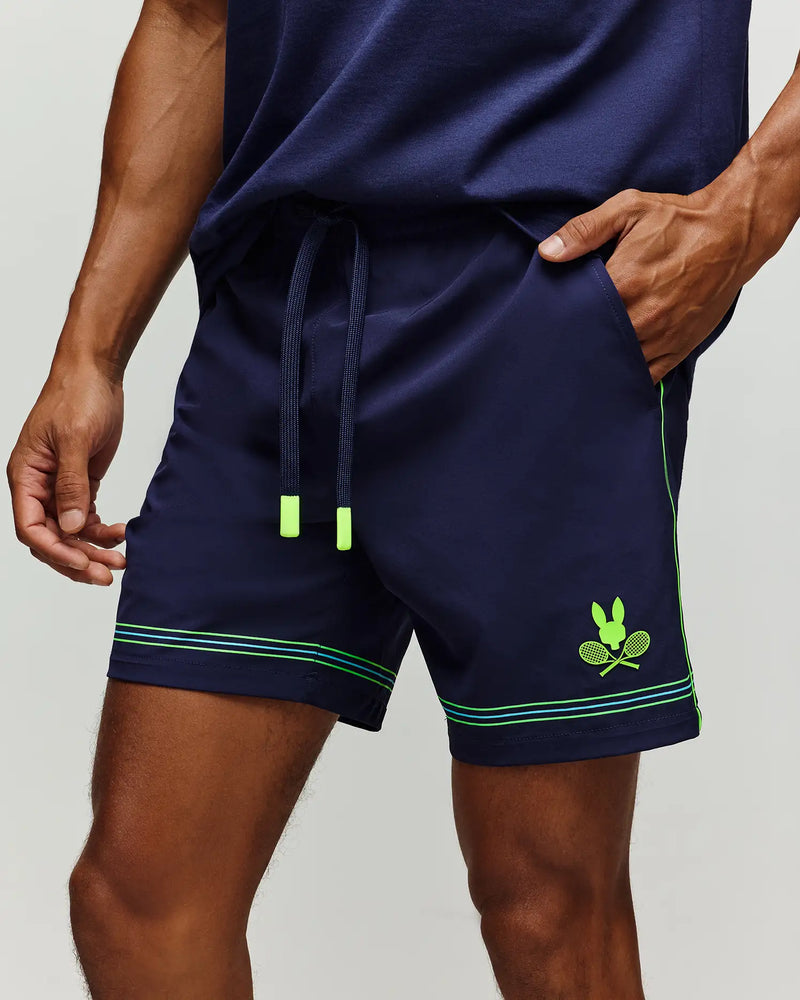 A person wears Psycho Bunny’s TENNIS BALL CREW MENS SHORT (MB0900017) in navy, featuring neon green accents, a drawstring, and a crossed rackets bunny logo graphic on the left leg. Their hand is in the right pocket.