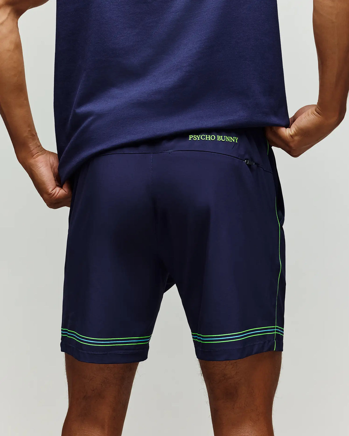 A man stands with hands on hips, back facing the camera, wearing Psycho Bunny TENNIS BALL CREW MEN'S SHORT (MB0900017) in navy blue with green stitching and "PSYCHO BUNNY" on the waistband—a sporty look ideal for tennis.