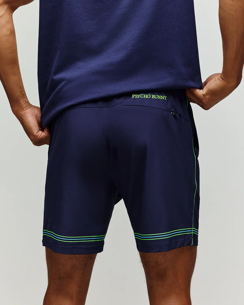 A man stands with hands on hips, back facing the camera, wearing Psycho Bunny TENNIS BALL CREW MEN'S SHORT (MB0900017) in navy blue with green stitching and "PSYCHO BUNNY" on the waistband—a sporty look ideal for tennis.
