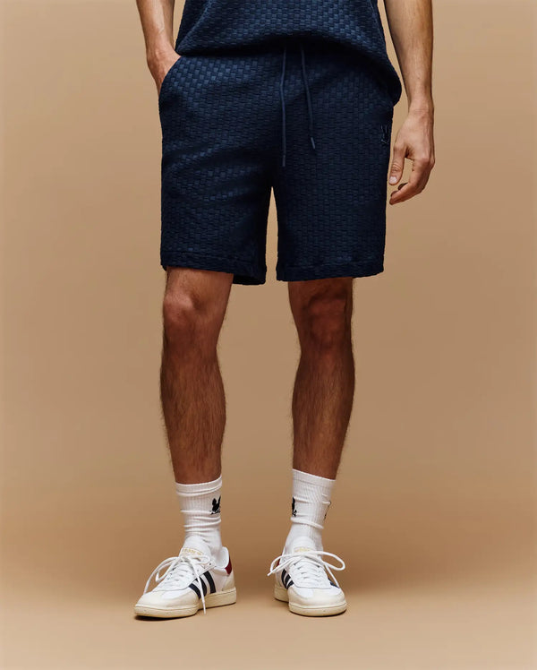 A person wears Psycho Bunny MENS KAIRO CROCHET SHORT - MB0900026 in navy, with a matching top, white socks featuring a black logo, and white sneakers. Only the lower torso and legs are seen against a beige background.