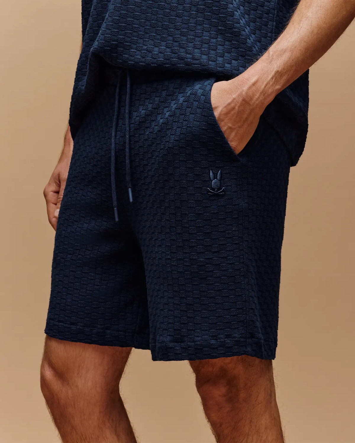 A person wears Psycho Bunny MENS KAIRO CROCHET SHORT - MB0900026 in textured navy blue premium cotton, hands in pockets, with a small embroidered logo on the left leg, standing against a beige background.