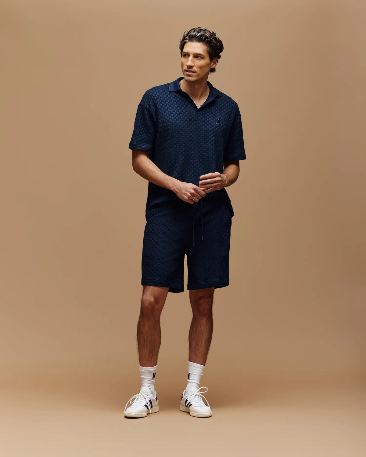 A man stands against a beige background wearing Psycho Bunny's MENS KAIRO CROCHET SHORT - MB0900026 in navy, paired with a matching collared shirt, white socks, and sneakers. He looks slightly to the side with hands loosely clasped.