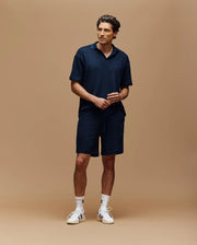 A man stands against a beige background wearing Psycho Bunny's MENS KAIRO CROCHET SHORT - MB0900026 in navy, paired with a matching collared shirt, white socks, and sneakers. He looks slightly to the side with hands loosely clasped.