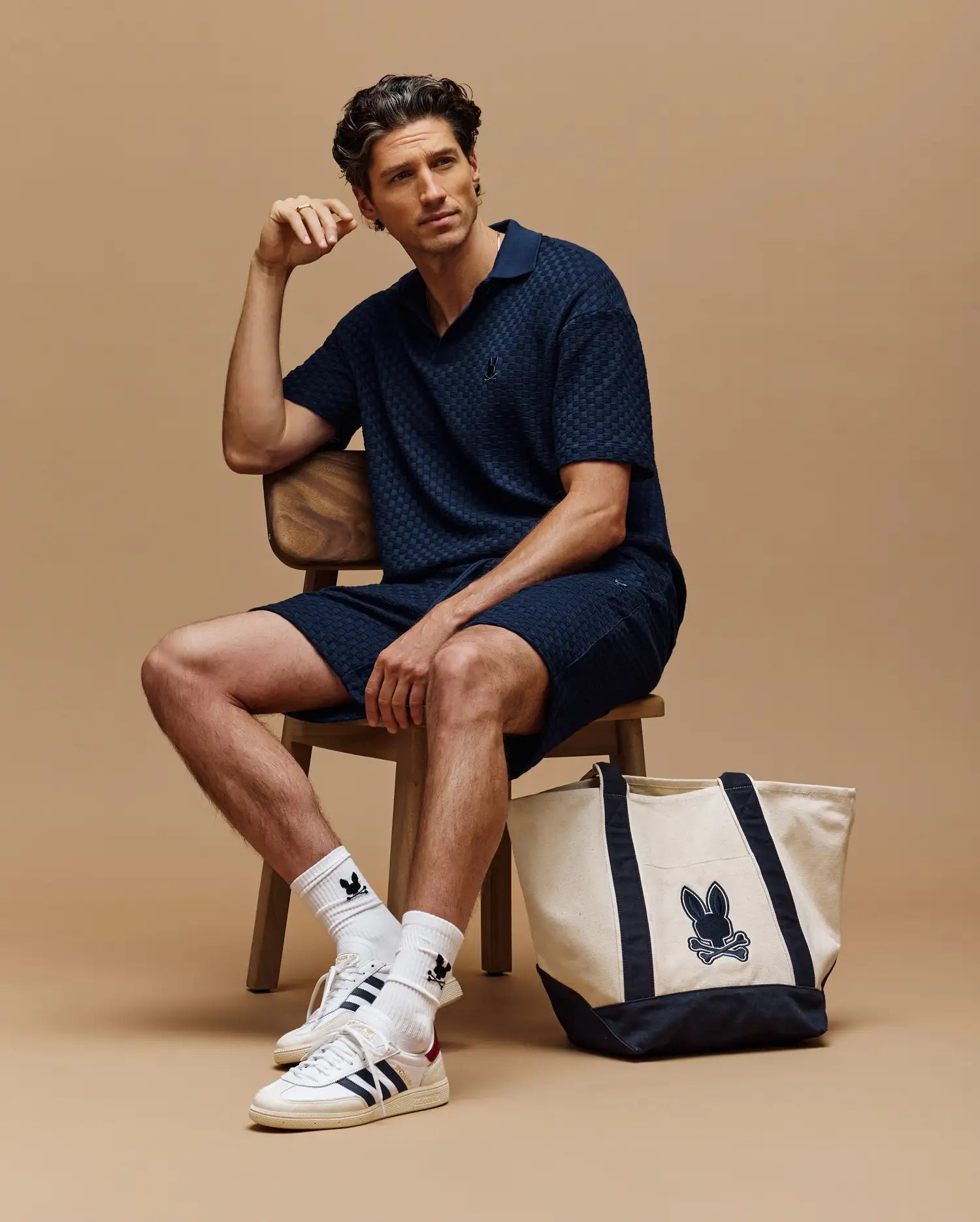 A man sits on a wooden stool against a beige background, wearing a navy blue polo, Psycho Bunny MENS KAIRO CROCHET SHORT - MB0900026, white socks and sneakers. A beige and navy Psycho Bunny tote with bunny logo is on the floor beside him.