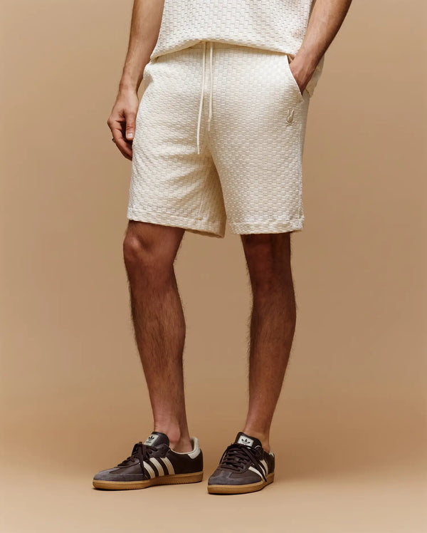 Against a beige background, only the lower body is visible, wearing Psycho Bunny MENS KAIRO CROCHET SHORT - MB0900026 in cream with a matching top and black Adidas sneakers with white stripes; hands relaxed by their sides—a perfect summer look.