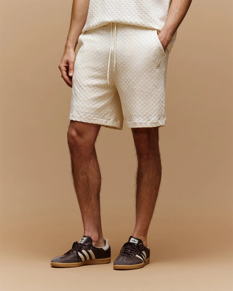 Against a beige background, only the lower body is visible, wearing Psycho Bunny MENS KAIRO CROCHET SHORT - MB0900026 in cream with a matching top and black Adidas sneakers with white stripes; hands relaxed by their sides—a perfect summer look.