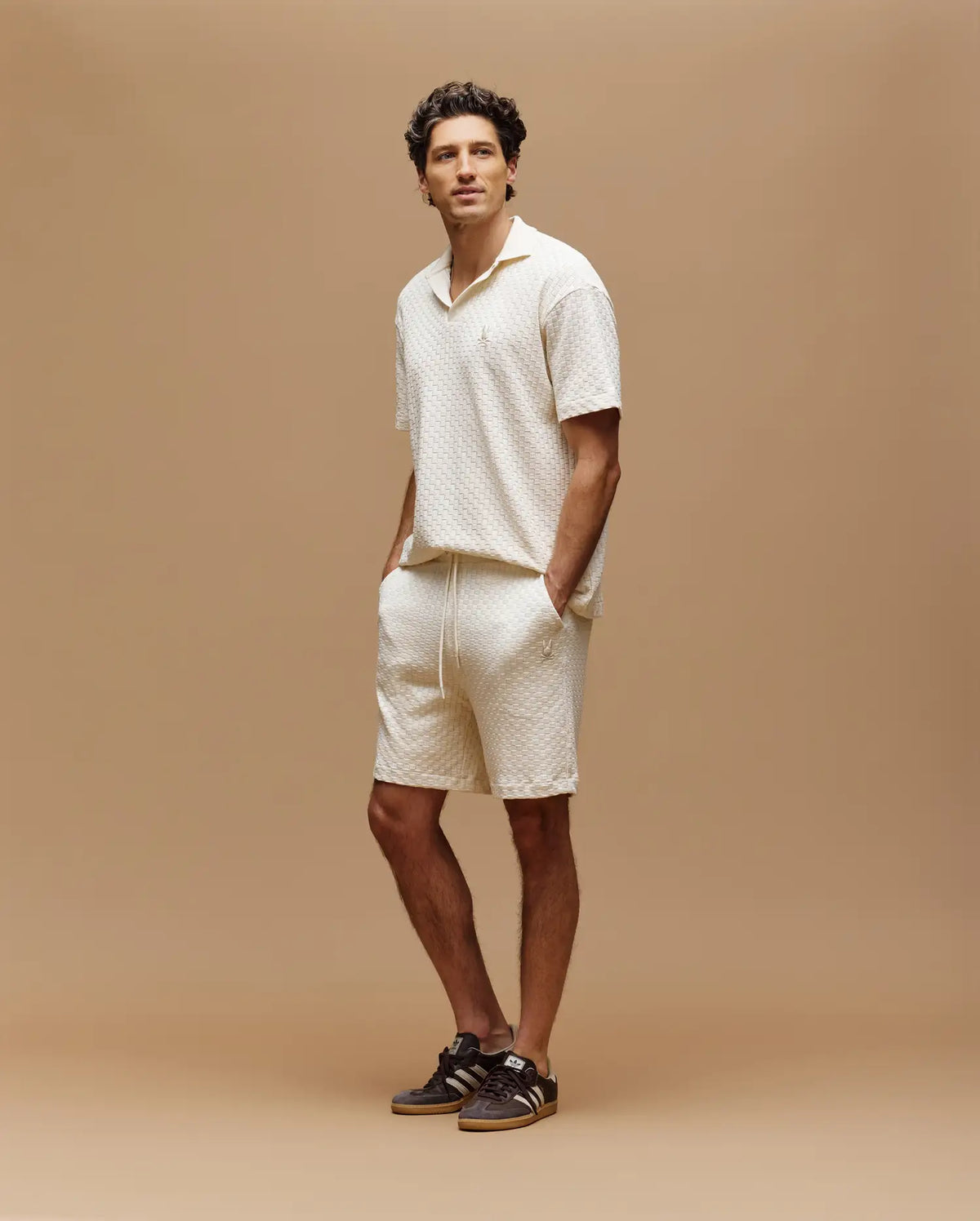 A man stands against a tan background, wearing Psycho Bunny's MENS KAIRO CROCHET SHORT - MB0900026 in cream with a short-sleeve shirt and brown sneakers. One hand is in his pocket.