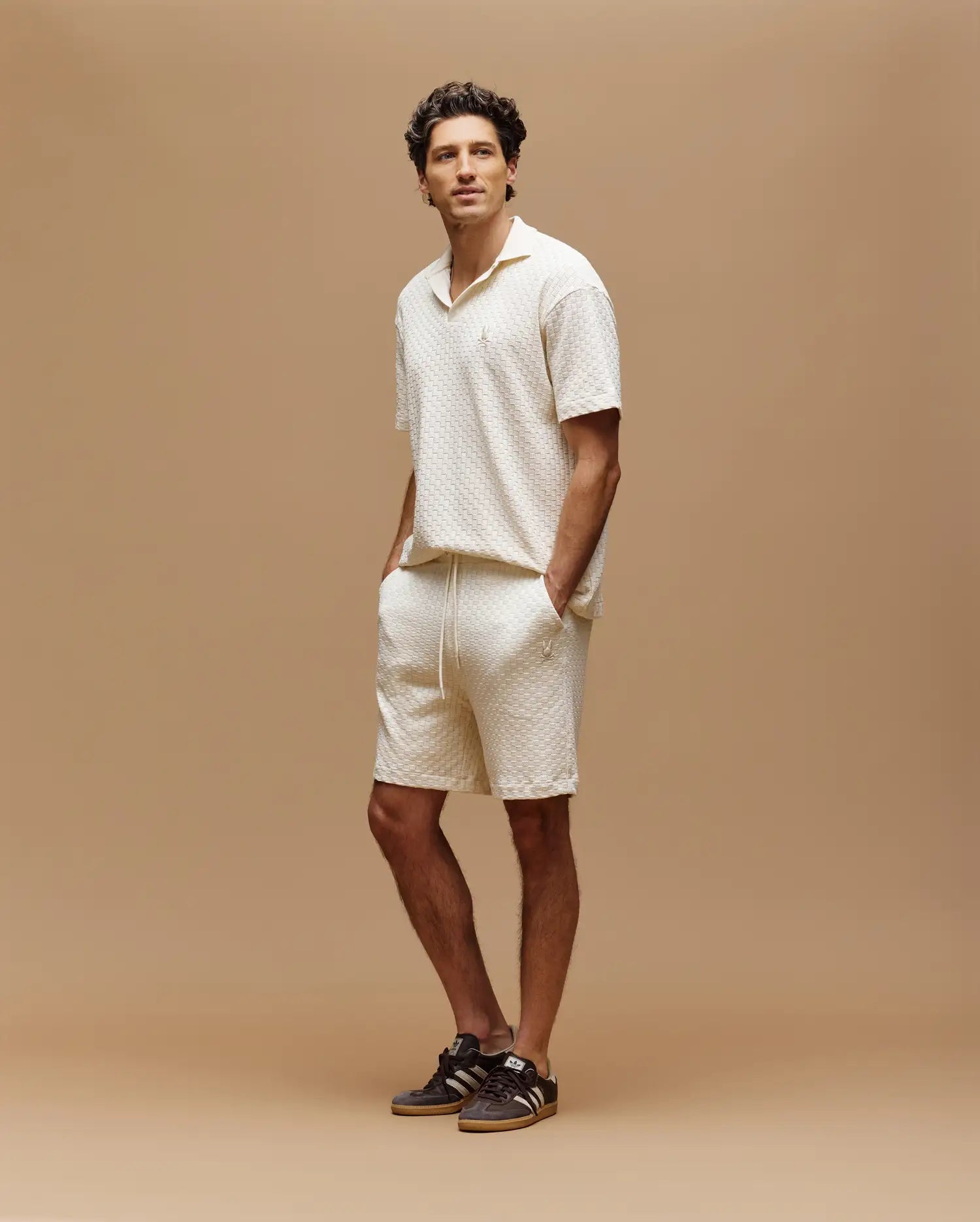 A man stands against a tan background, wearing Psycho Bunny's MENS KAIRO CROCHET SHORT - MB0900026 in cream with a short-sleeve shirt and brown sneakers. One hand is in his pocket.