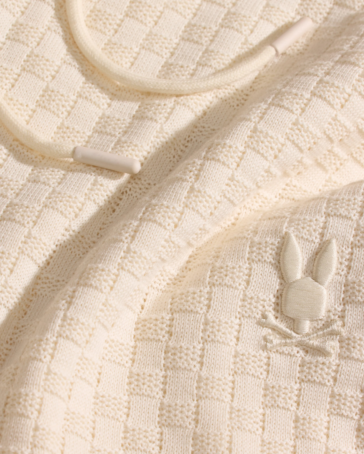 Close-up of Psycho Bunny’s MENS KAIRO CROCHET SHORT (MB0900026) in cream-colored, checkered knit with a raised bunny and crossbones logo. Features a cream drawstring with plastic tips—ideal for summer wear.