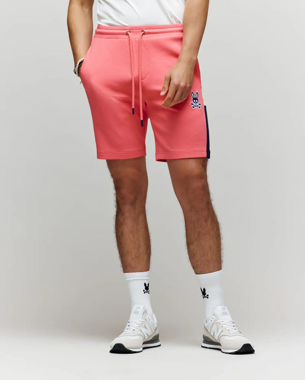 A man showcases modern street style in Psycho Bunny's pink MENS DAWSON TRACK SHORT - MB0900032 with a skull bunny logo, paired with a white tee, white sneakers, and white socks featuring a black bunny logo, against a plain light background.