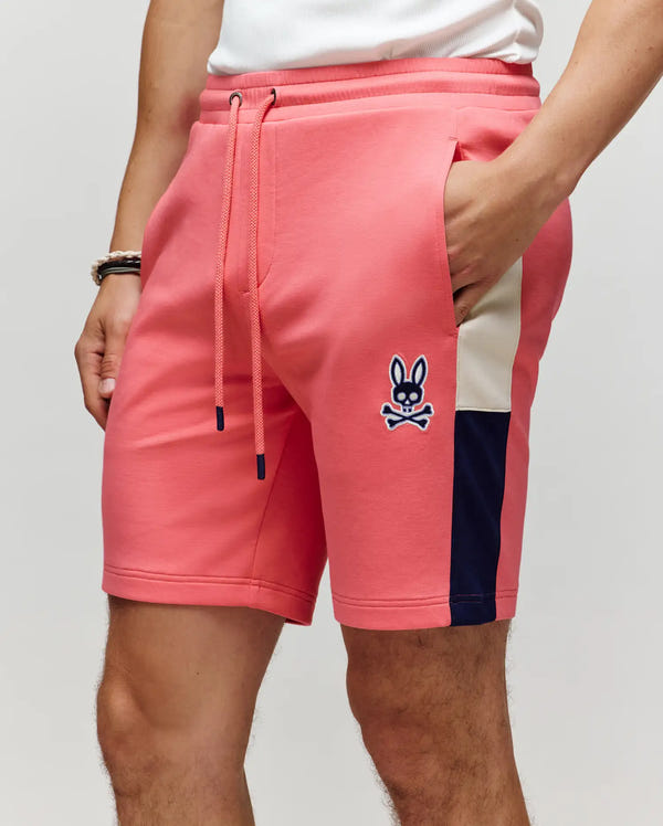 A person wears Psycho Bunny MENS DAWSON TRACK SHORT - MB0900032 in coral pink, with a drawstring, white and navy side stripe, and blue bunny skull-and-crossbones logo by the pocket. Hand is in the pocket for a relaxed street style look.