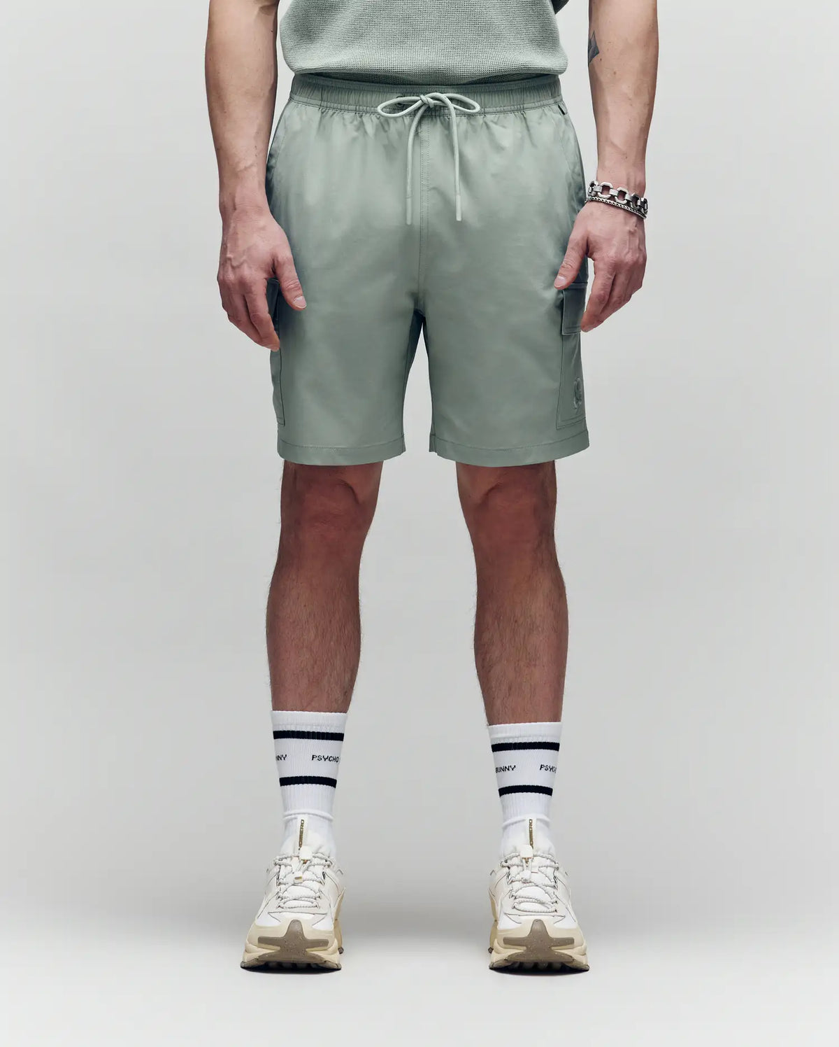 A person stands against a plain background wearing Psycho Bunny MENS RANDY BOARD SWIM SHORT - MB1000002 in sage green with cargo pockets, a coordinating shirt, white striped socks, cream sneakers, and a silver bracelet. The image is from mid-torso to feet.