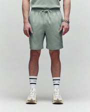 A person stands against a plain background wearing Psycho Bunny MENS RANDY BOARD SWIM SHORT - MB1000002 in sage green with cargo pockets, a coordinating shirt, white striped socks, cream sneakers, and a silver bracelet. The image is from mid-torso to feet.